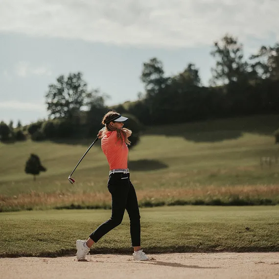 Golferin schwingt Golfschläger auf einem grünen Feld, Bäume im Hintergrund.