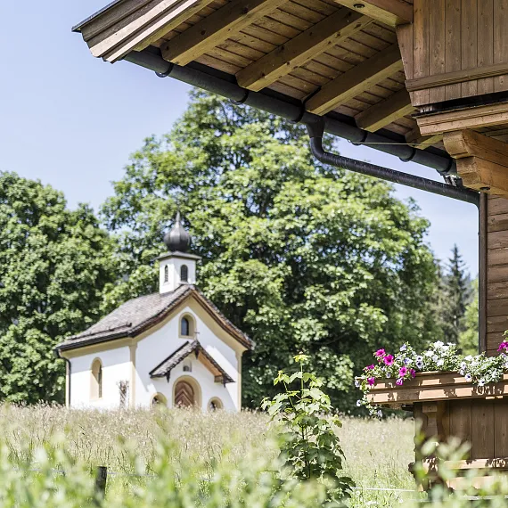 Kleine Kapelle in einer grünen Landschaft mit Bäumen, im Vordergrund Holzhausdach mit Blumen.