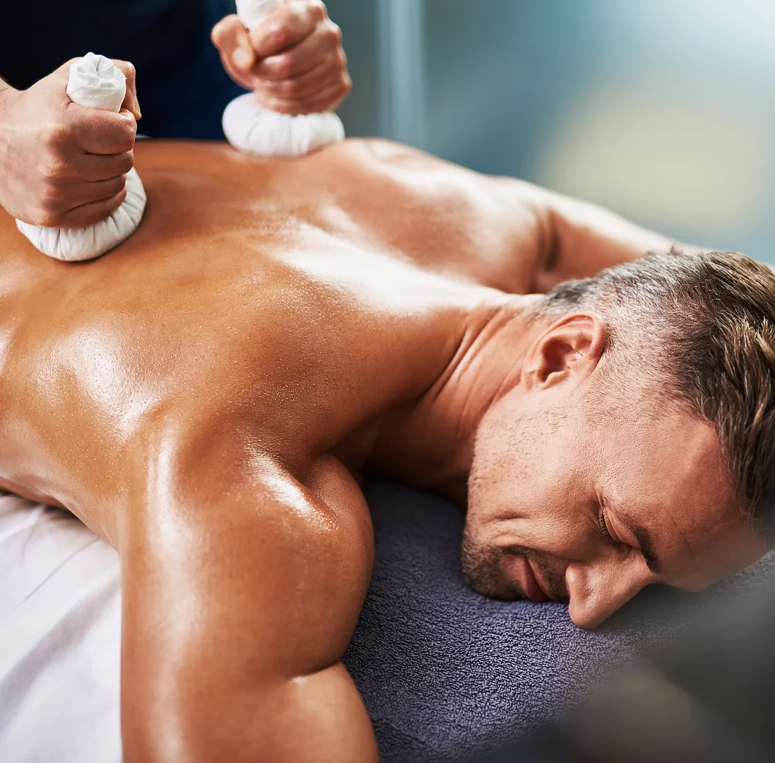 Mann liegt entspannt auf Massagebank, Rücken wird mit Kräuterstempeln massiert, entspannte Wellness-Umgebung.
