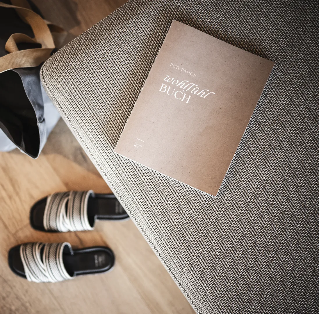 Notizbuch auf grauem Sessel, daneben Sandalen und eine Tasche auf Holzfußboden.