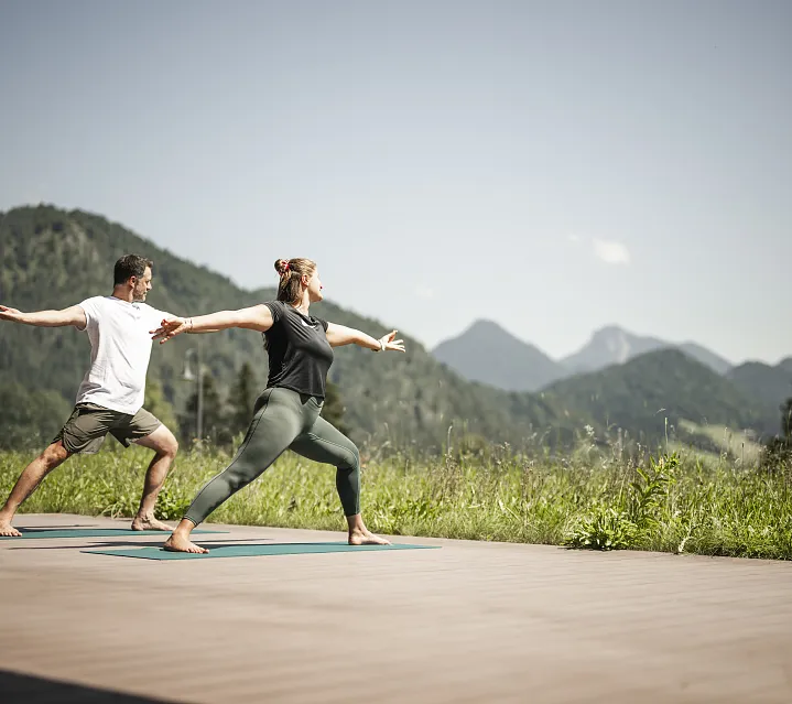 Zwei Personen praktizieren Yoga auf einer Plattform mit Blick auf Tiroler Berge.