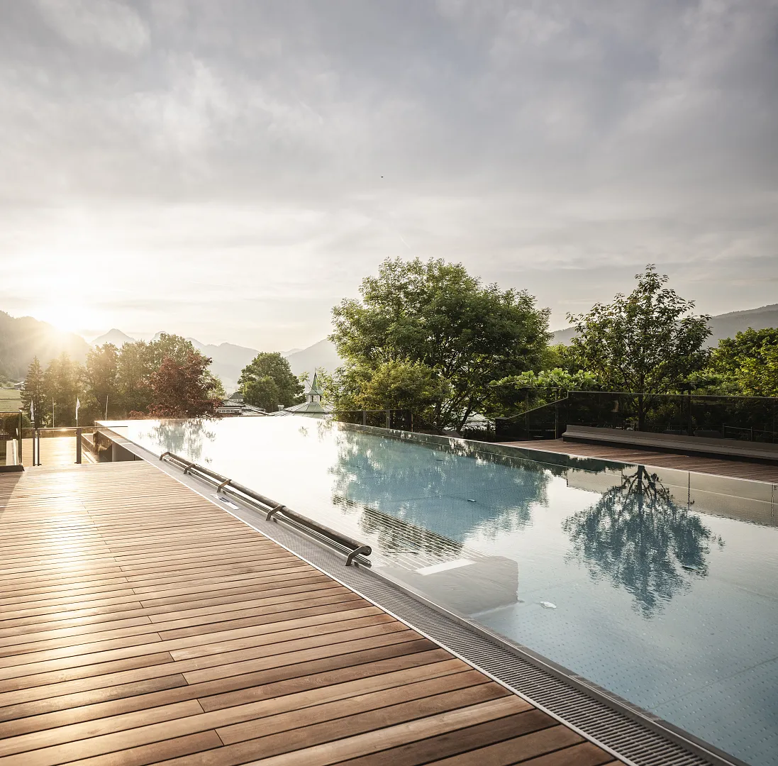 Infinity-Pool mit Holzterrasse, umgeben von Bäumen, in einer bergigen Landschaft bei Sonnenuntergang.