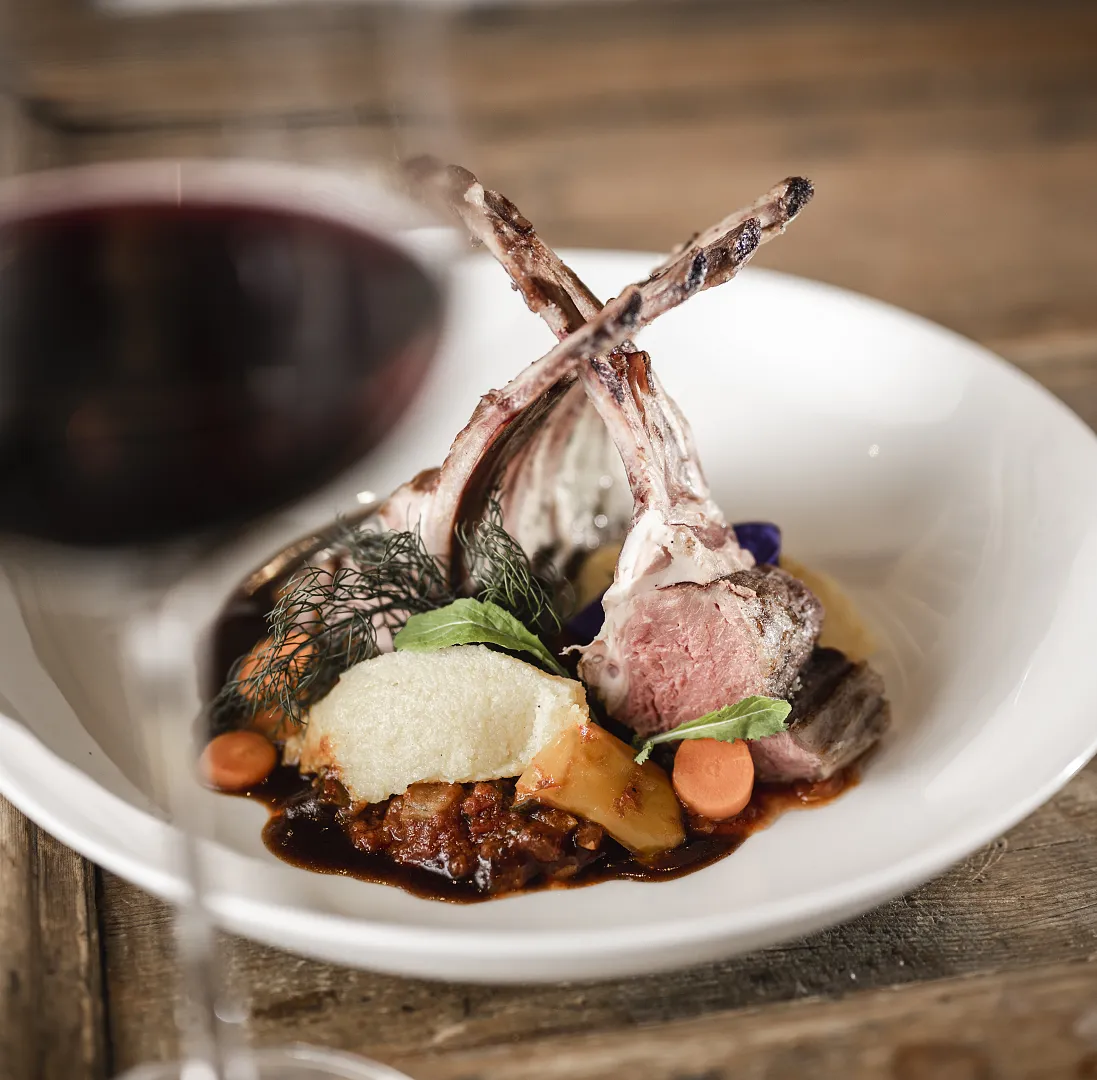 Plate with roasted lamb chops, vegetables, and sauce; glass of red wine in the foreground.