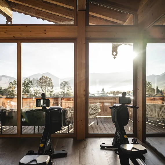 Blick aus einem modernen Fitnessraum auf eine sonnige Berglandschaft in Tirol, mit Fitnessgeräten im Vordergrund.