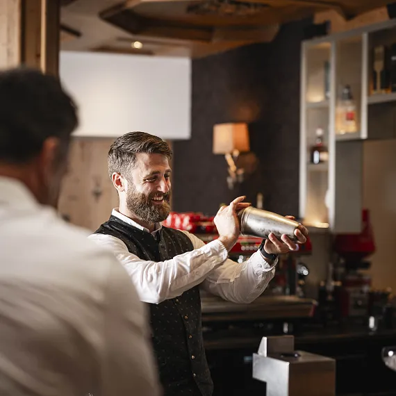 Barkeeper mixt einen Cocktail in einer Bar mit dunkler Einrichtung, eine andere Person im Vordergrund.