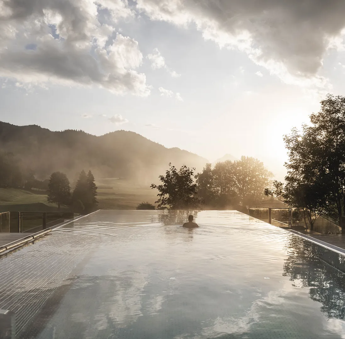 Unendlicher Pool im Freien mit Alpenkulisse bei Sonnenuntergang, umgeben von Bäumen und Bergen in Tirol.