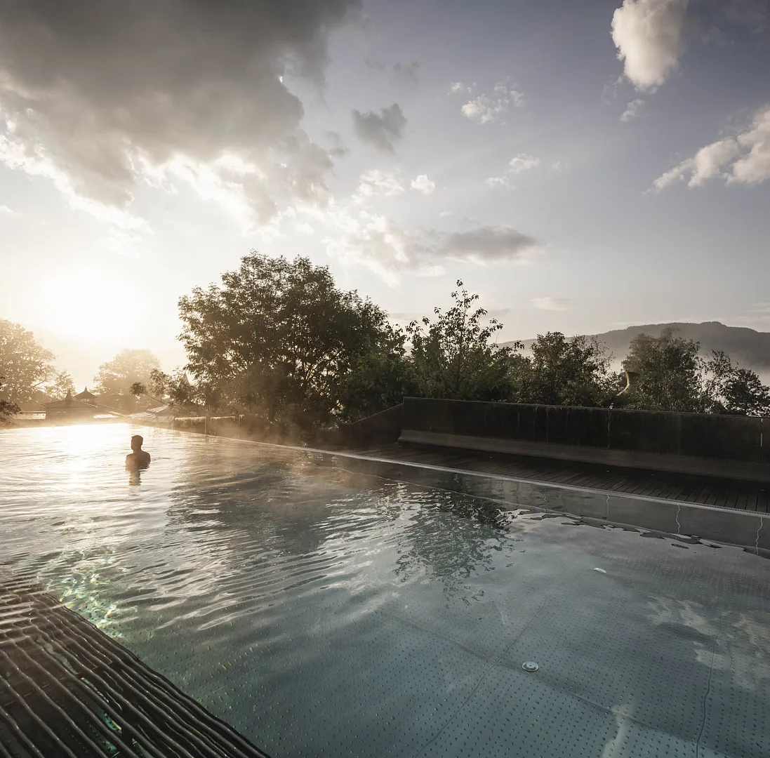 Person schwimmt in einem ruhigen Außenpool bei Sonnenuntergang, umgeben von Bäumen und Bergen.