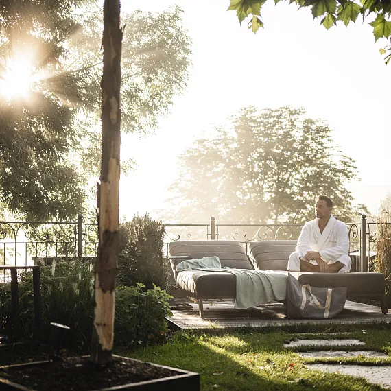 Person im Bademantel auf Sofa in Garten bei Sonnenaufgang, umgeben von Bäumen.