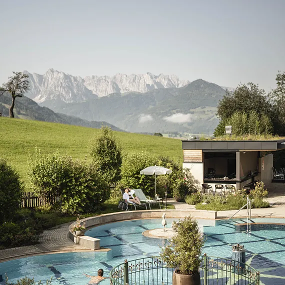 Zwei Personen entspannen auf Liegestühlen am Pool vor Bergkulisse in Tirol.