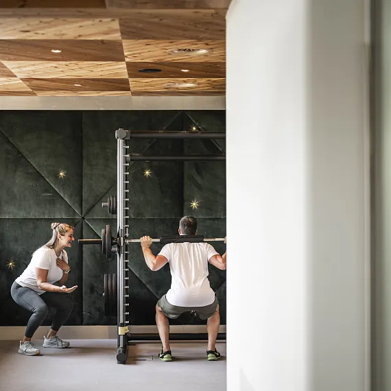 Person hebt Langhantel in einem modernen Fitnessstudio, zweite Person assistiert nebenher.
