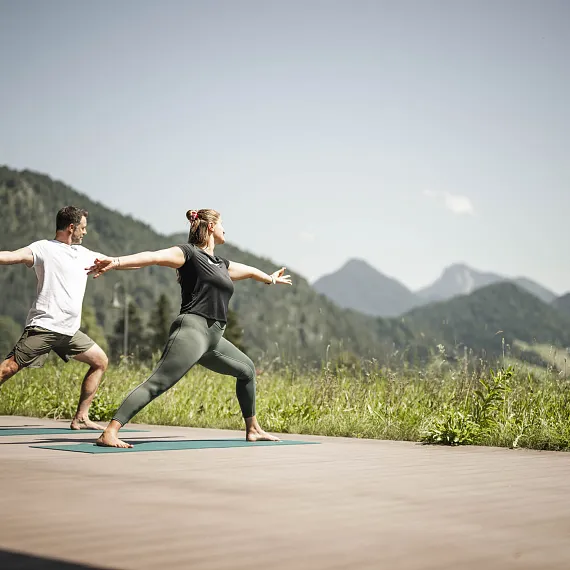 Zwei Personen praktizieren Yoga auf einer Terrasse vor der Bergkulisse von Kitzbühel, Tirol.