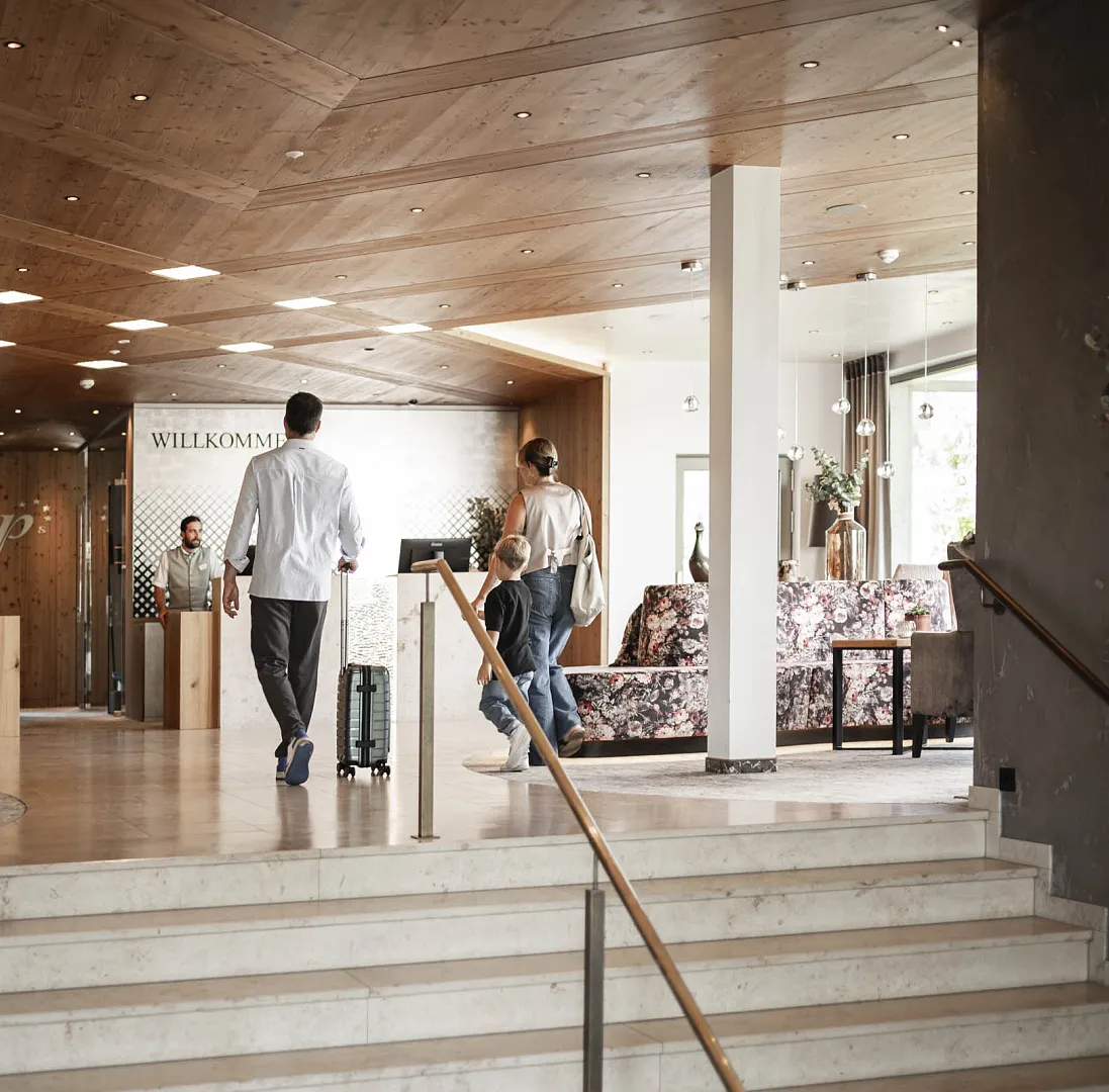 Personen betreten ein edel gestaltetes Hotel-Foyer mit Gepäck am Empfangsbereich.