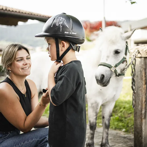 Eine Frau hilft einem Kind, einen Reithelm anzulegen, während ein weißes Pony daneben steht.