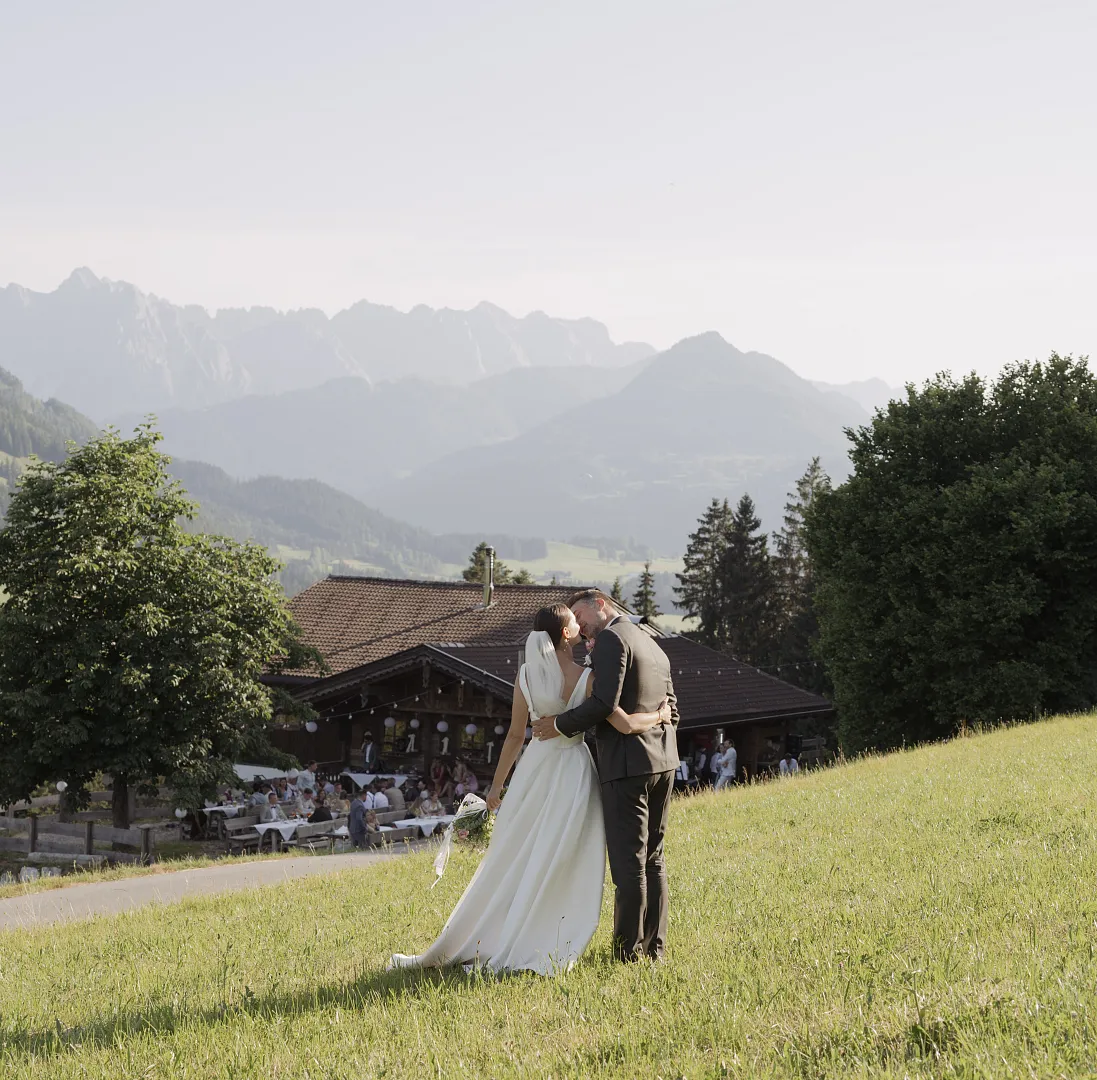Brautpaar küsst sich auf einer Wiese in den Tiroler Bergen, eine Hütte und Berglandschaft im Hintergrund.