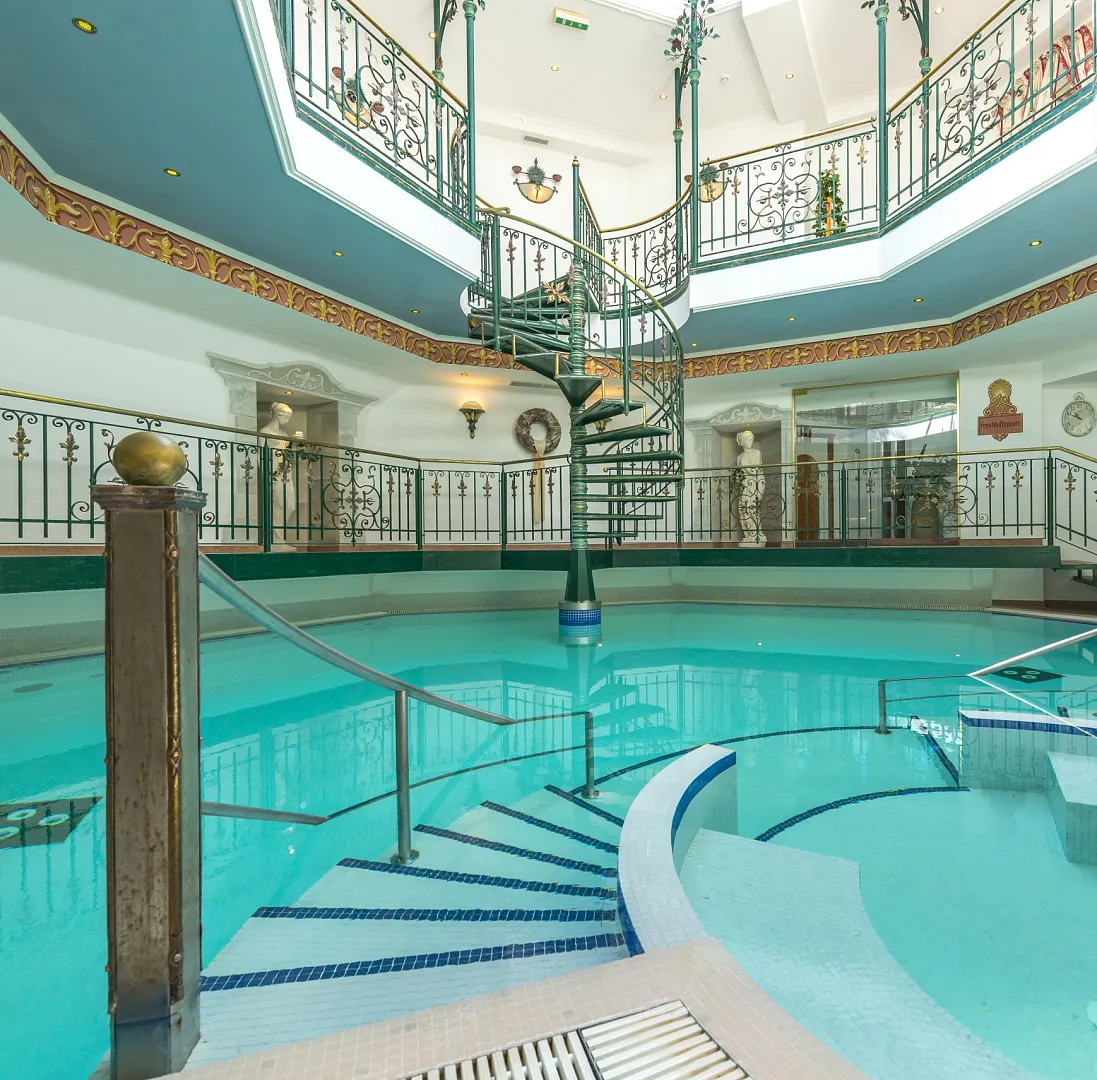 Innenraum eines Hotels mit rundem Pool und Wendeltreppe, umgeben von Geländern und dekorativen Elementen.