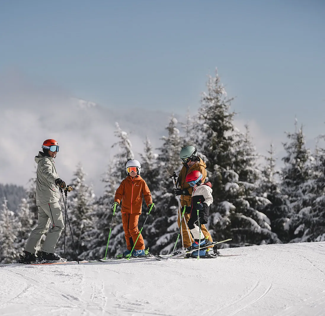 Erwachsener und drei Kinder in Skiausrüstung auf Skipiste, umgeben von verschneiten Bäumen, sonniger Tag.
