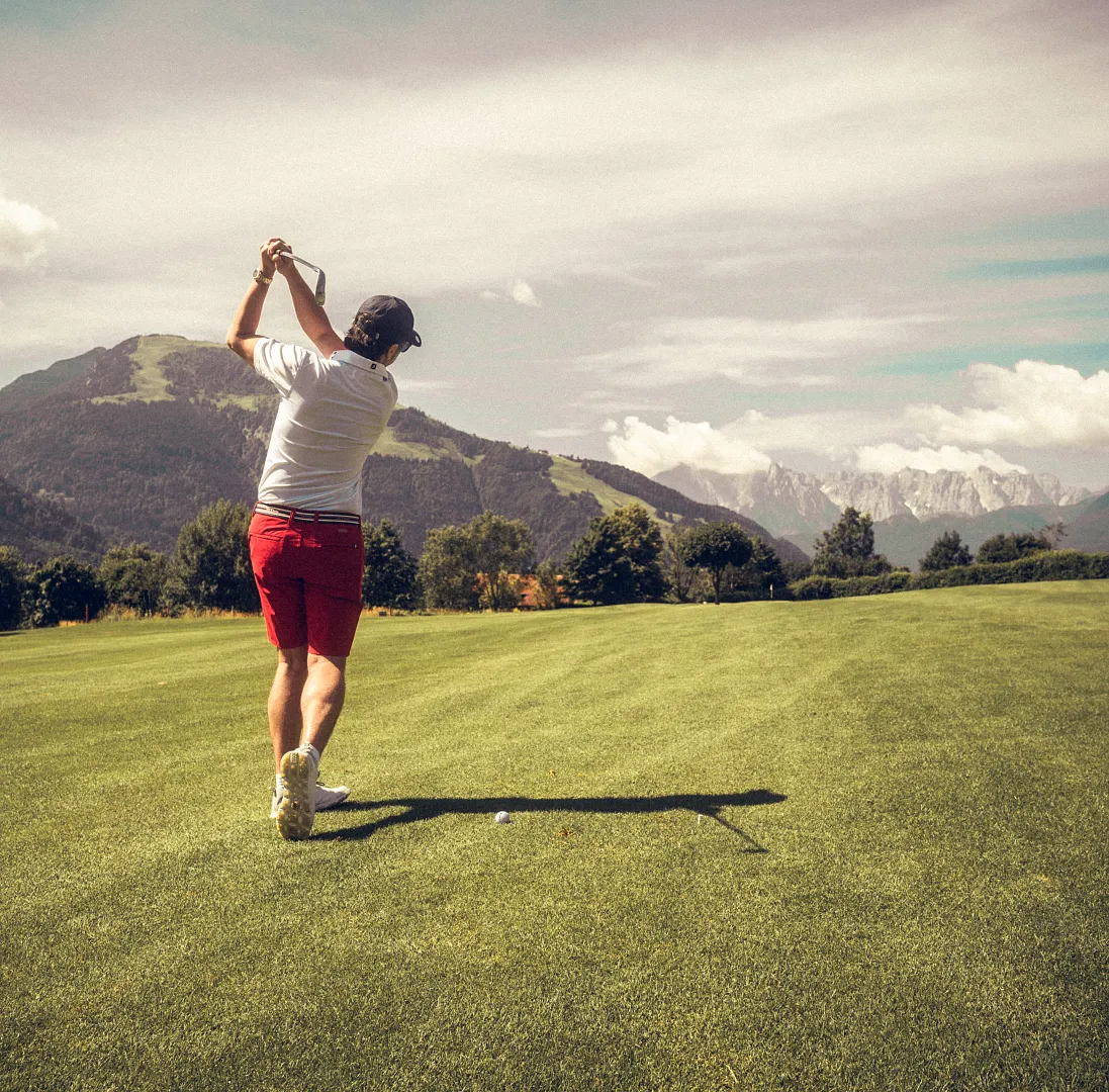 Person schwingt Golfschläger auf einem grünen Golfplatz, umgeben von Bergen und Wolken.
