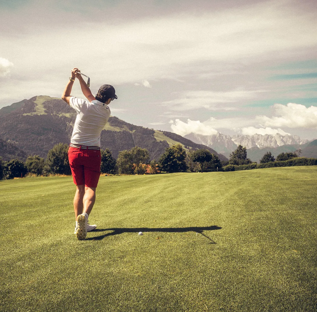 Person schwingt Golfschläger auf einem grünen Golfplatz, umgeben von Bergen und Wolken.