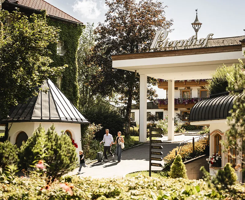 Zwei Erwachsene und ein Kind gehen durch einen gepflegten Garten mit traditionellen Gebäuden im Hintergrund.