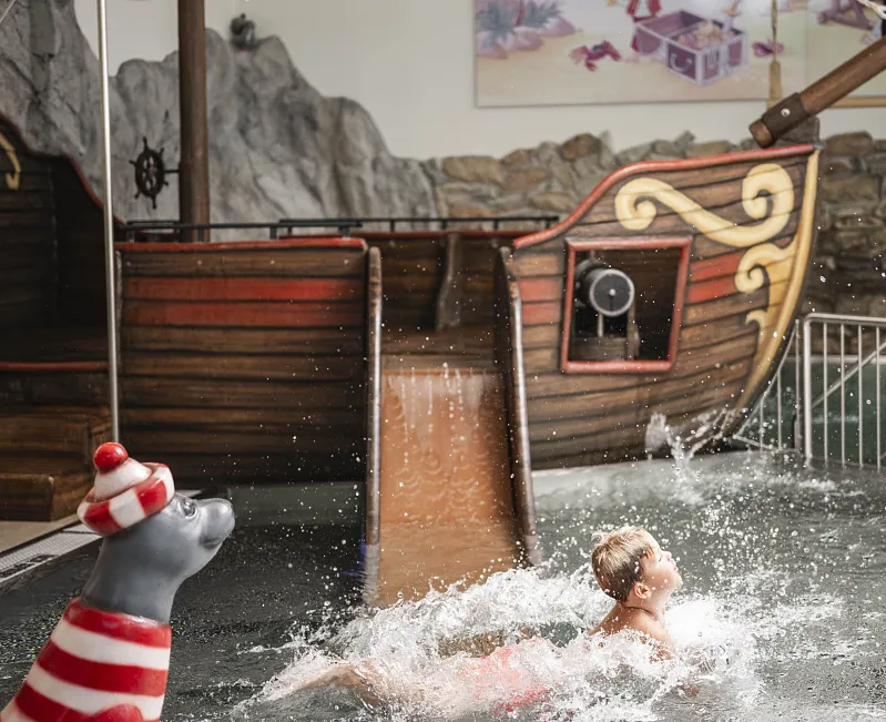 Kind plätschert im Wasser vor einem Piratenschiff-Spielplatz mit Rutsche.