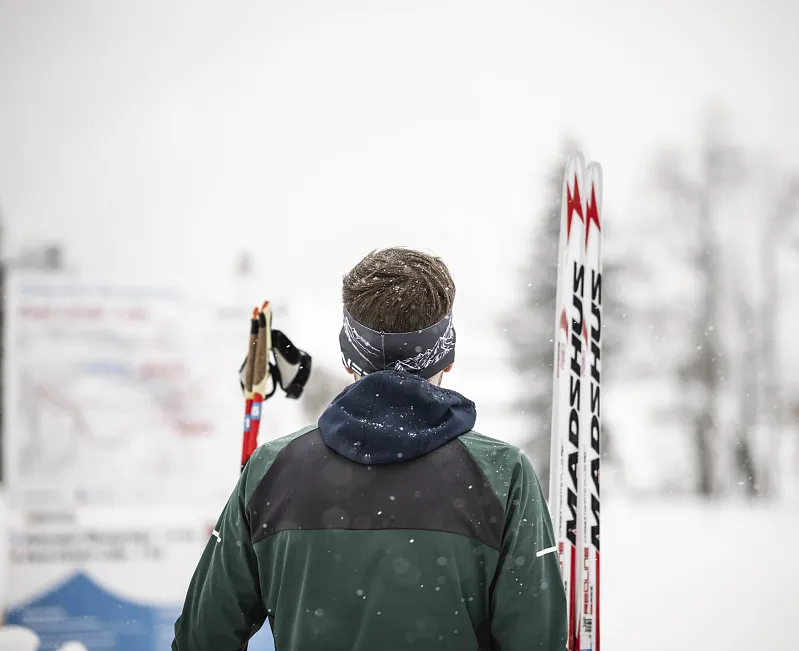Person mit Skiern schaut auf eine verschneite Informationstafel, hinter der Schnee liegt.