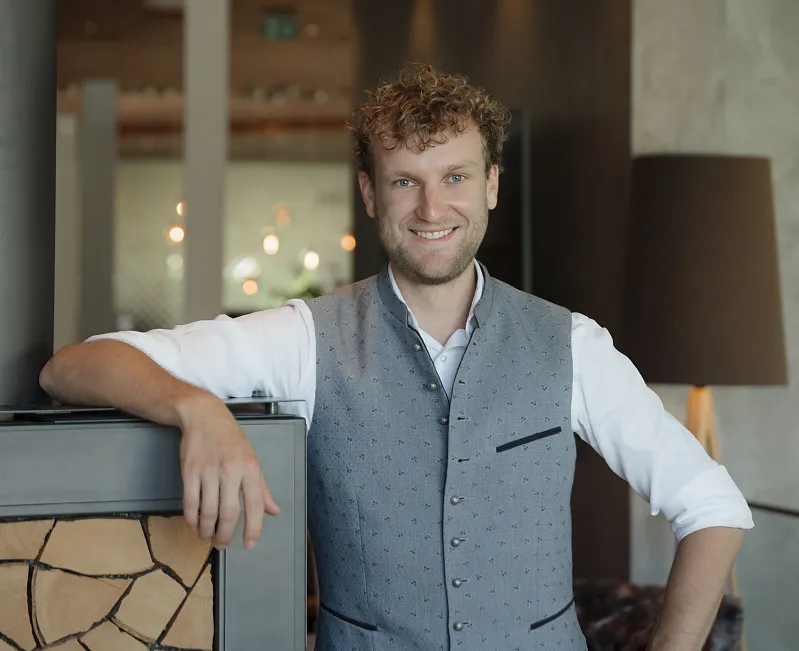 Ein Mann in traditioneller, eleganter Weste steht lächelnd neben einem modernen Innenkamin.
