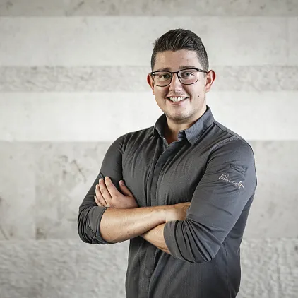Person mit Brille und dunklem Hemd steht vor einer grauen Wand, lächelt mit verschränkten Armen.