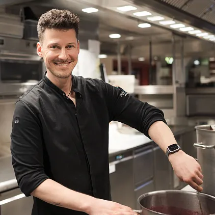 Koch in professioneller Küche mit großem Topf, lächelnd, trägt schwarze Kleidung und Smartwatch.