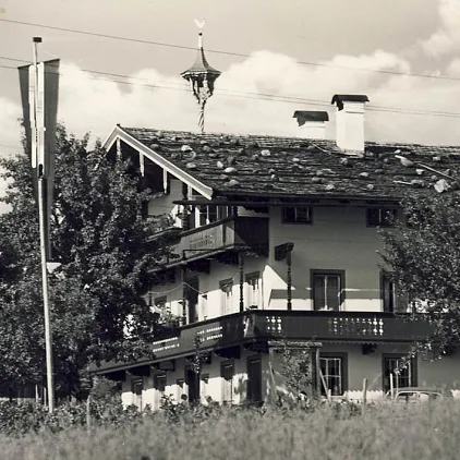 Traditionelles Haus mit Holzbalkon, Fahne und Bäumen in ländlicher Umgebung, möglicherweise in Tirol.