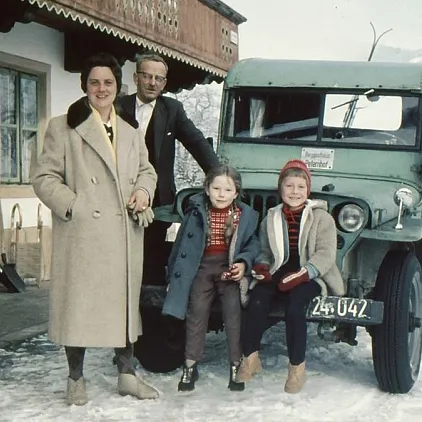 Gruppe von vier Personen posiert im Winter vor einem grünen Fahrzeug vor einem Haus mit Skiern an der Wand.