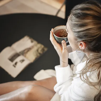 Frau in Bademantel trinkt Kaffee und liest ein Buch auf einem Tisch liegend.
