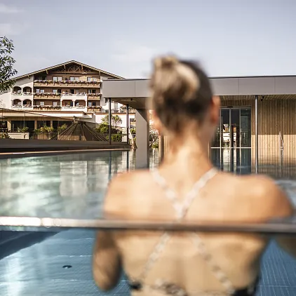 Frau steht im Infinity-Pool mit Blick auf modernes Hotelgebäude und Alpenkulisse.