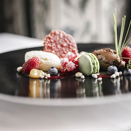 Elegantes Dessert mit Macaron, Mousse, Beeren und dekorativen Elementen auf einem schwarzen Teller.