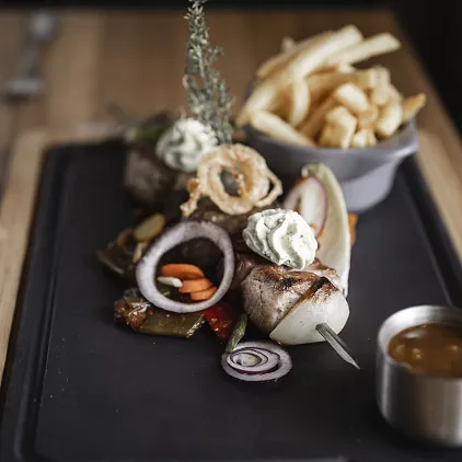 Holzbrett mit gegrilltem Fleischspieß, Zwiebelringen, Kräuterbutter und Pommes im Hintergrund.