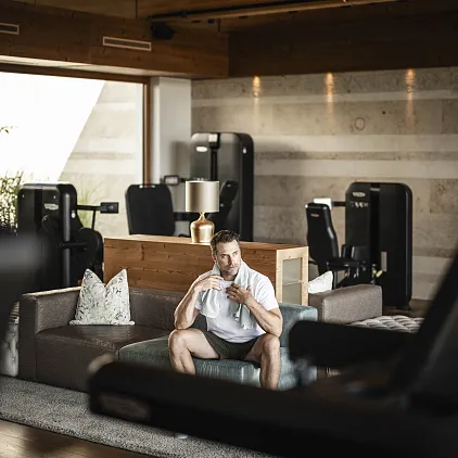 Mann im modernen Fitnessstudio mit Holzausstattung, umgeben von Trainingsgeräten, sitzt auf einer Ledercouch.