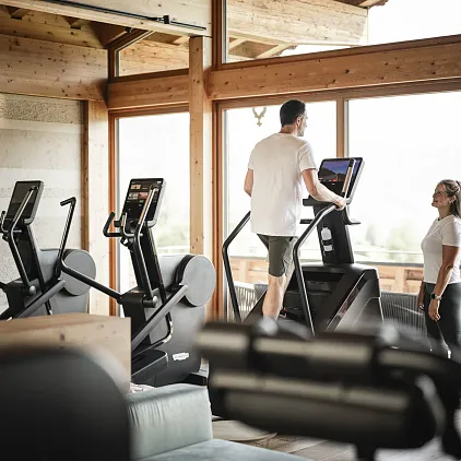 Mann auf Laufband und Frau daneben in einem Fitnessstudio mit Holzinterieur und großen Fenstern.