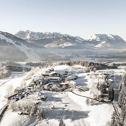 Luftaufnahme einer verschneiten Landschaft in Tirol mit Bergen im Hintergrund und Siedlung im Vordergrund.
