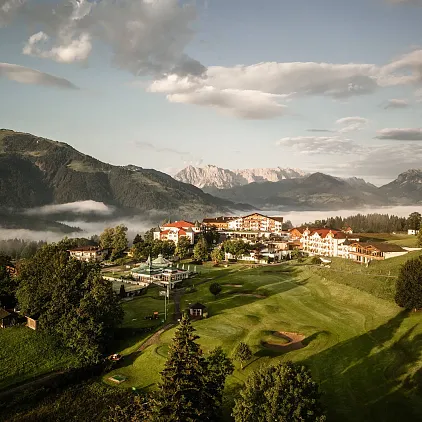 Landschaft in Kitzbühel, Tirol, mit Bergen im Hintergrund und einem Golfplatz im Vordergrund unter bewölktem Himmel.