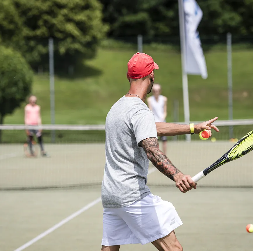 Mann mit rotem Hut spielt Tennis auf einem Außenplatz.