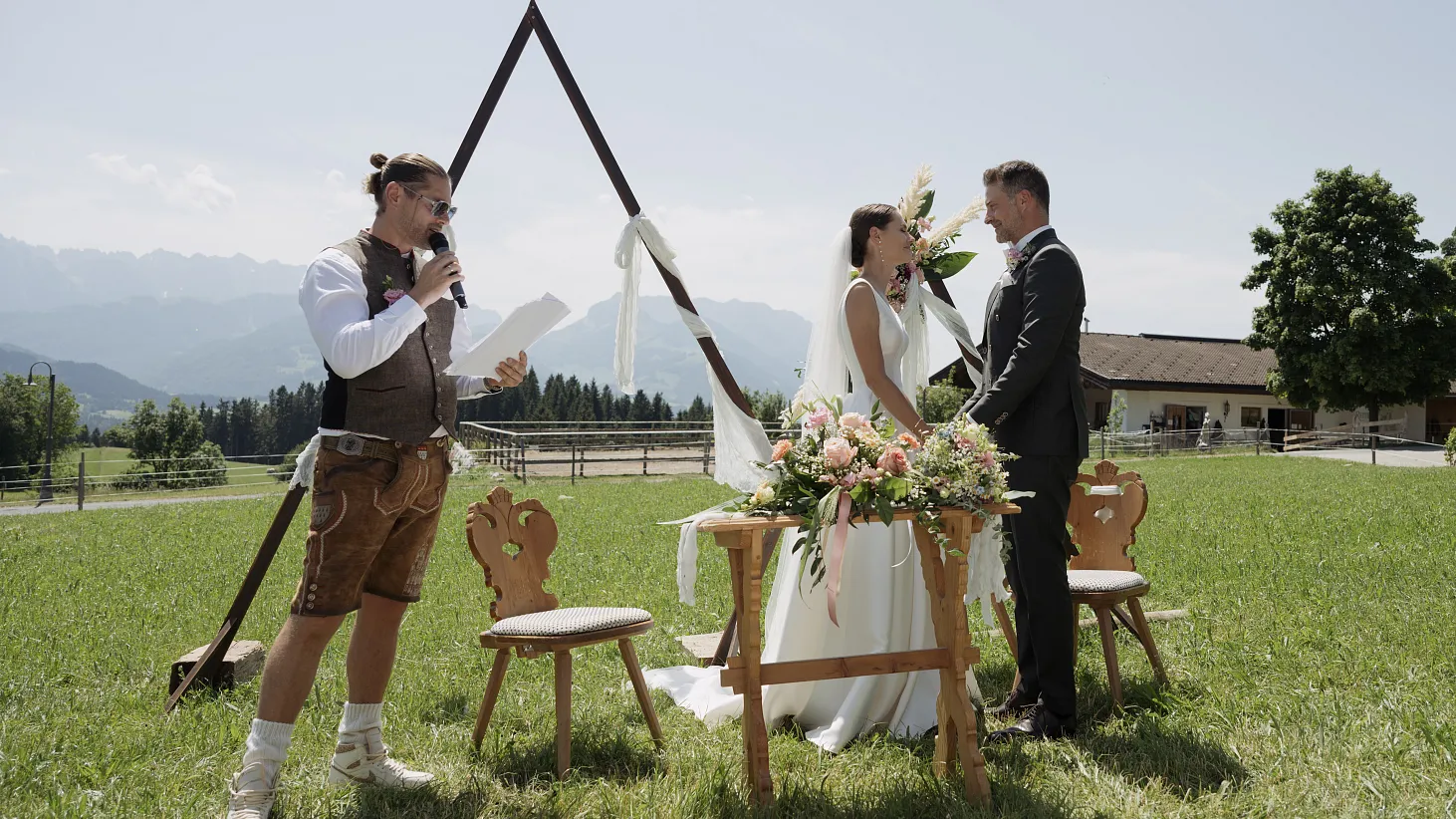 A wedding ceremony outdoors with a couple and officiant in traditional attire, set in a scenic mountainous landscape.