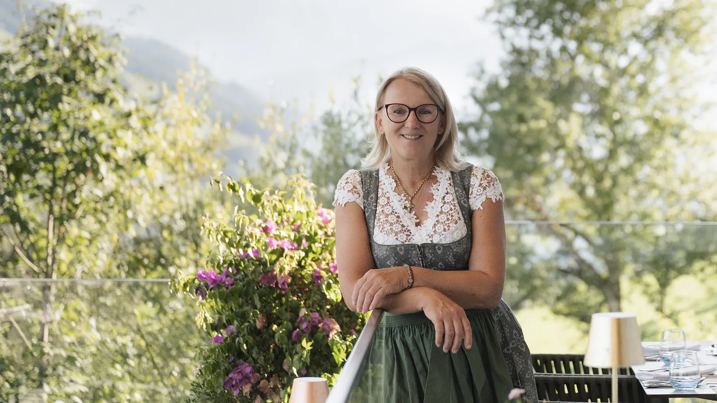 Frau in traditioneller Tracht lehnt an einem Geländer, umgeben von Bäumen und Bergen in Tirol.