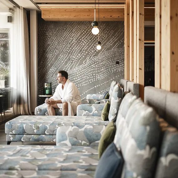 Person in a white robe seated in a modern lounge with patterned wall and blue sofas.