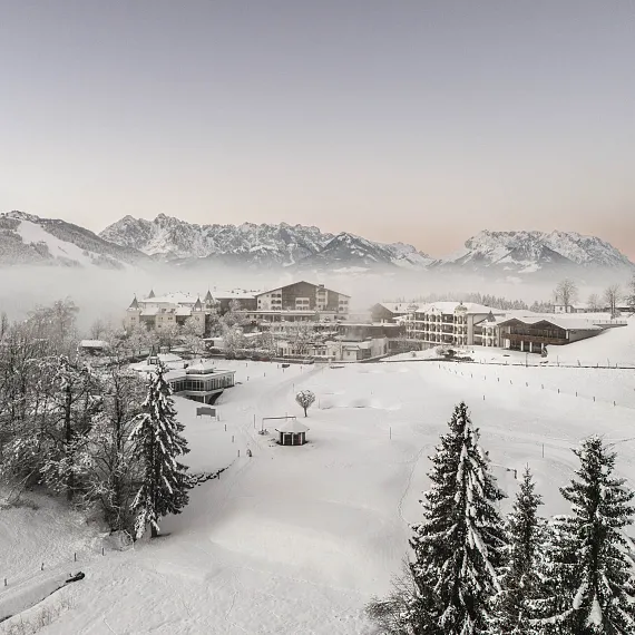 Verschneite winterliche Landschaft in Kitzbühel, Tirol, mit Bergen im Hintergrund und dichten Wäldern im Vordergrund.