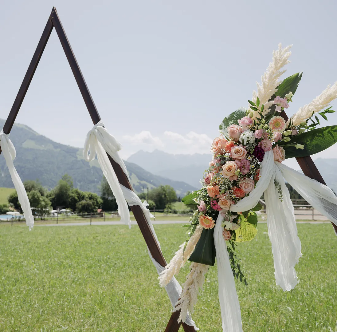 Dreieckiger Hochzeitsbogen mit Blumengesteck auf einer Wiese in den Bergen.