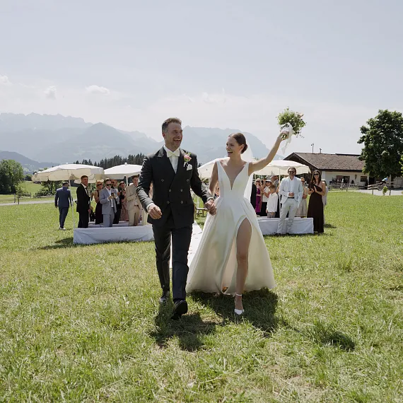 Bride and groom walking on a grassy field with guests seated behind, mountains in the background.