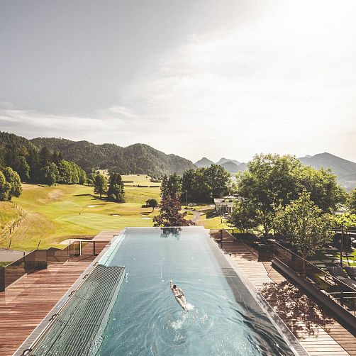 Person schwimmt in einem Infinity-Pool mit Blick auf grüne Hügel und Wälder in Kitzbühel, Tirol.