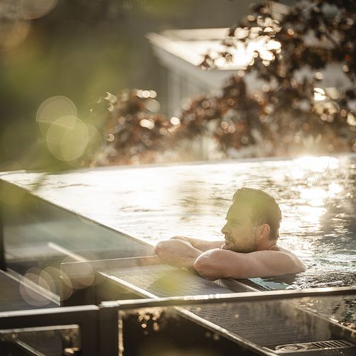 Mann entspannt im Infinity-Pool mit Naturkulisse im Hintergrund bei Sonnenuntergang.