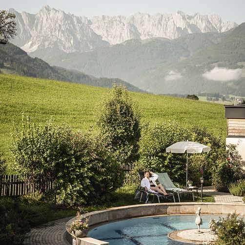 Mensch in Liegestuhl auf einer Wiese vor Bergkulisse in Tirol, unter einem Sonnenschirm sitzend.