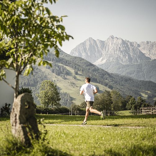 Person joggt auf einer Wiese vor einer Berglandschaft in Tirol bei klarem Wetter.