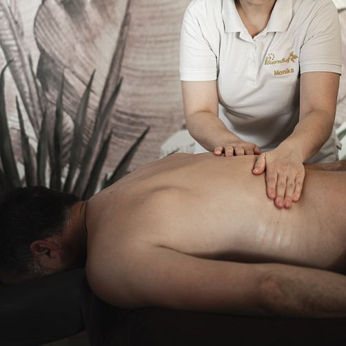 Eine Person in weißem Polohemd massiert den Rücken einer liegenden Person vor einer Blättertapete.